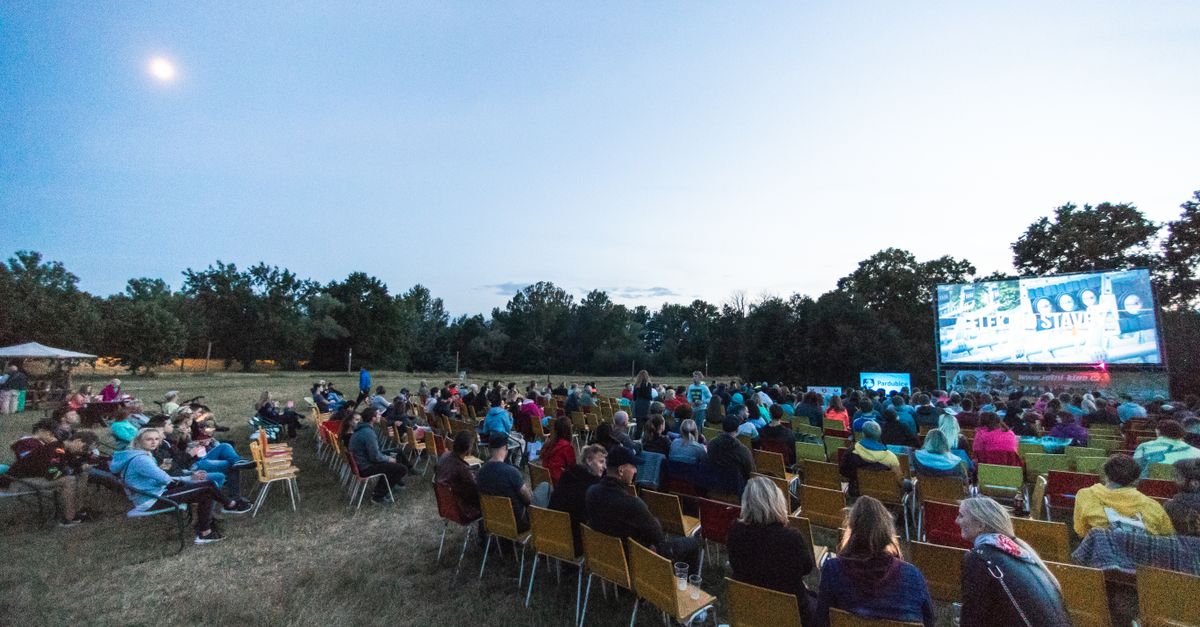 A large group enjoys a movie outdoors at night in a field with a big screen under the moon.